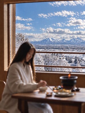 Eine Person sitzt an einem Tisch mit Blick auf schneebedeckte Berge und einen klaren Himmel.