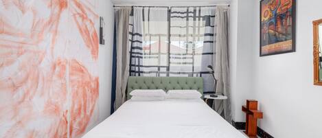 A bedroom with a bed, a green headboard, a painting, and a window with curtains.