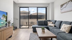 Ein modernes Wohnzimmer mit einem grauen Sofa, einem Holztisch und einem Fernseher auf einer Holzkommode. Ein großes Schiebeglasfenster führt auf einen Balkon mit Blick auf Gebäude und einen klaren Himmel.