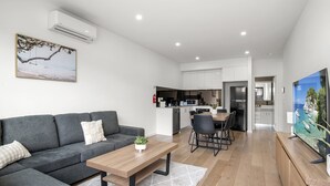 Ein modernes Wohnzimmer mit einer grauen Eckcouch, einem Couchtisch aus Holz und einem Essbereich mit Tisch und Stühlen. Im Hintergrund ist eine Küche mit weißen Schränken und einem Kühlschrank zu sehen.