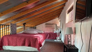 A bedroom with a sloped ceiling, a bed with a red and white checkered bedspread, a standing fan, and a small table with a lamp.