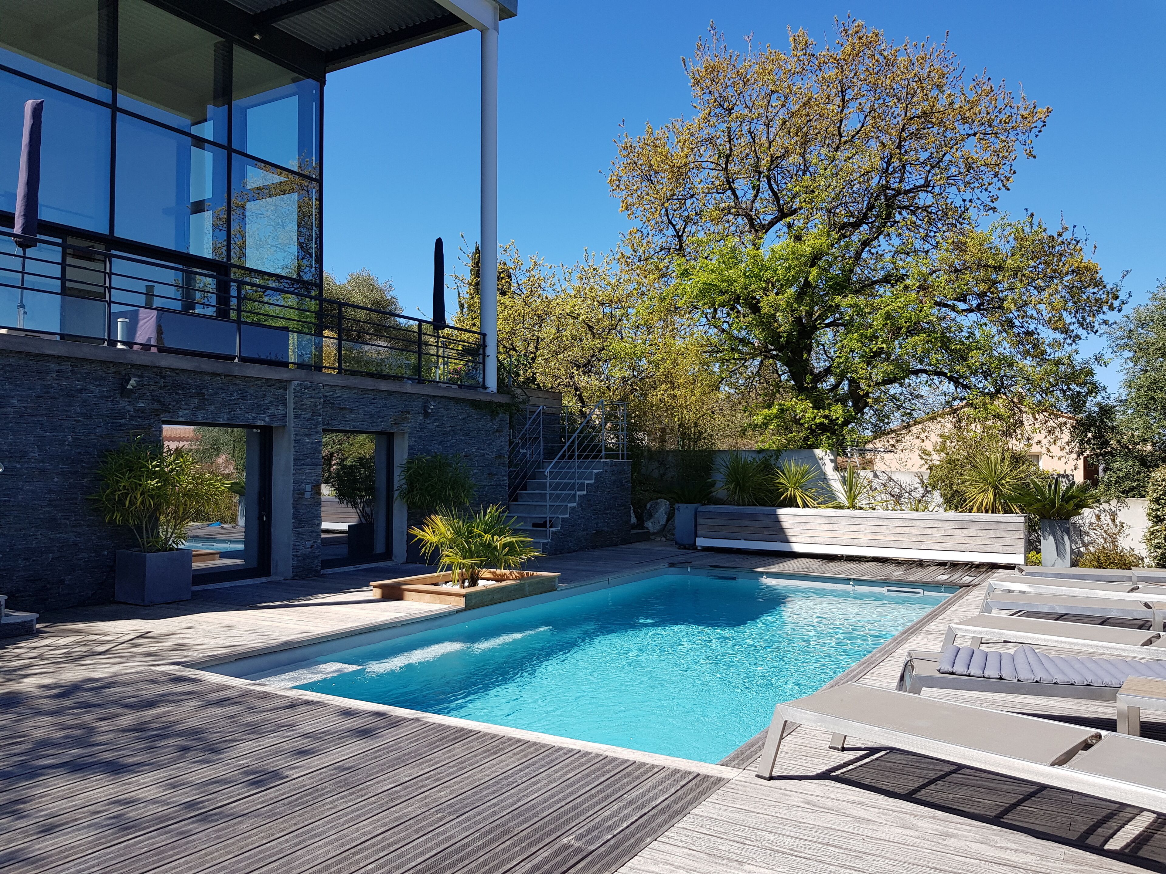 Zona de piscina moderna al aire libre con terraza de madera, sillas de descanso y un árbol grande.