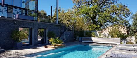 Zona de piscina moderna al aire libre con terraza de madera, sillas de descanso y un árbol grande.