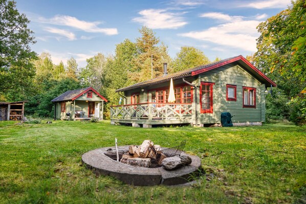 A rustic cabin with a wooden deck and a fire pit in the yard. - Hus PIO – Your Lakeside Holiday Retreat in Southern Sweden (Traryd)