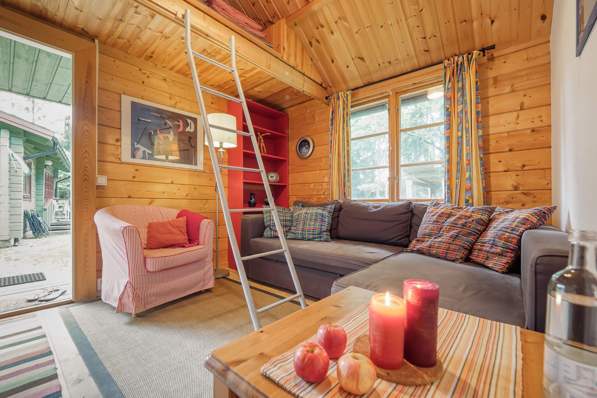 Un interior acogedor de cabaña con detalles de madera, un sofá, una silla, una mesa con manzanas y velas, y una escalera.