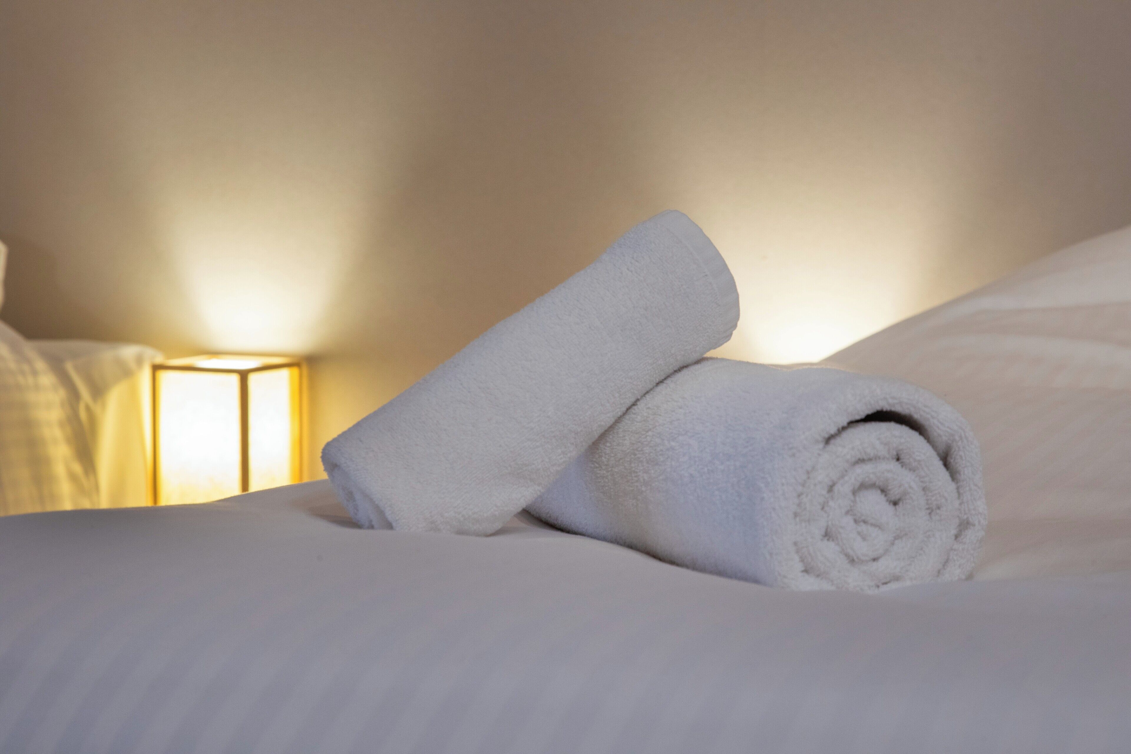 A neatly made bed with rolled white towels and a bedside lamp.
