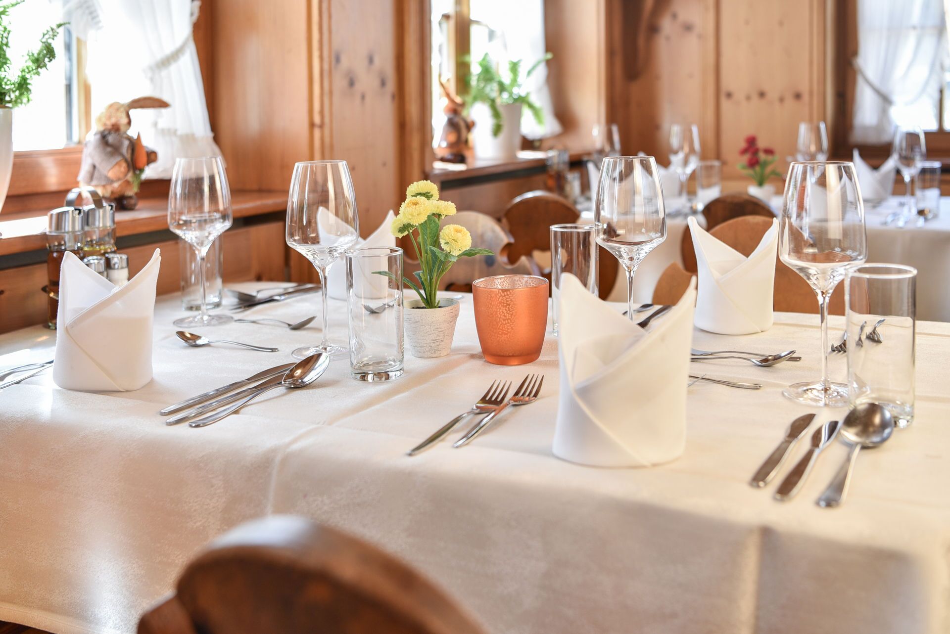 Una mesa de comedor dispuesta con varias copas de vino, tenedores y cucharas, una pequeña planta en maceta y un centro de mesa decorativo de vidrio.