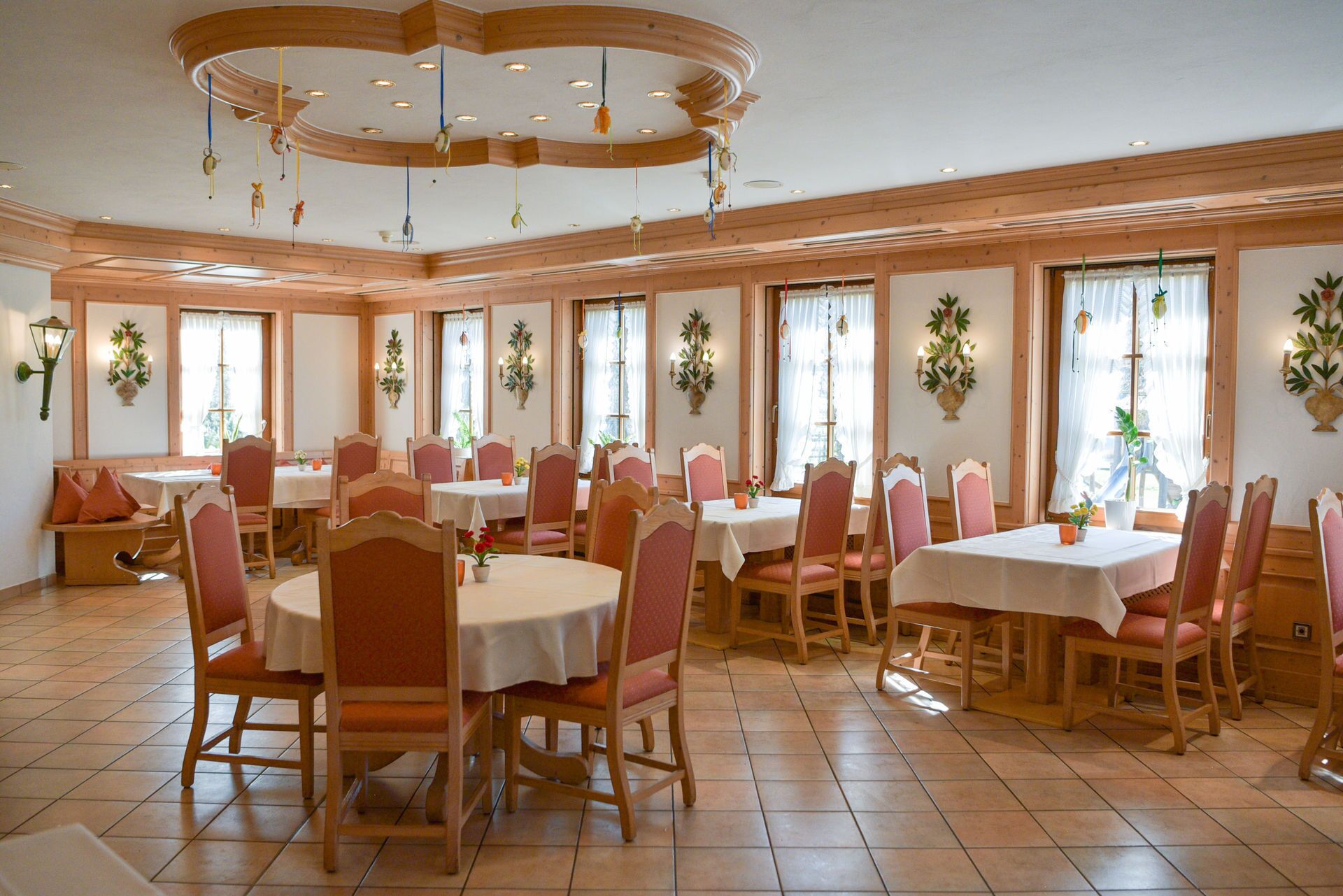A dining area with round tables and chairs, wooden paneling, and large windows with curtains.