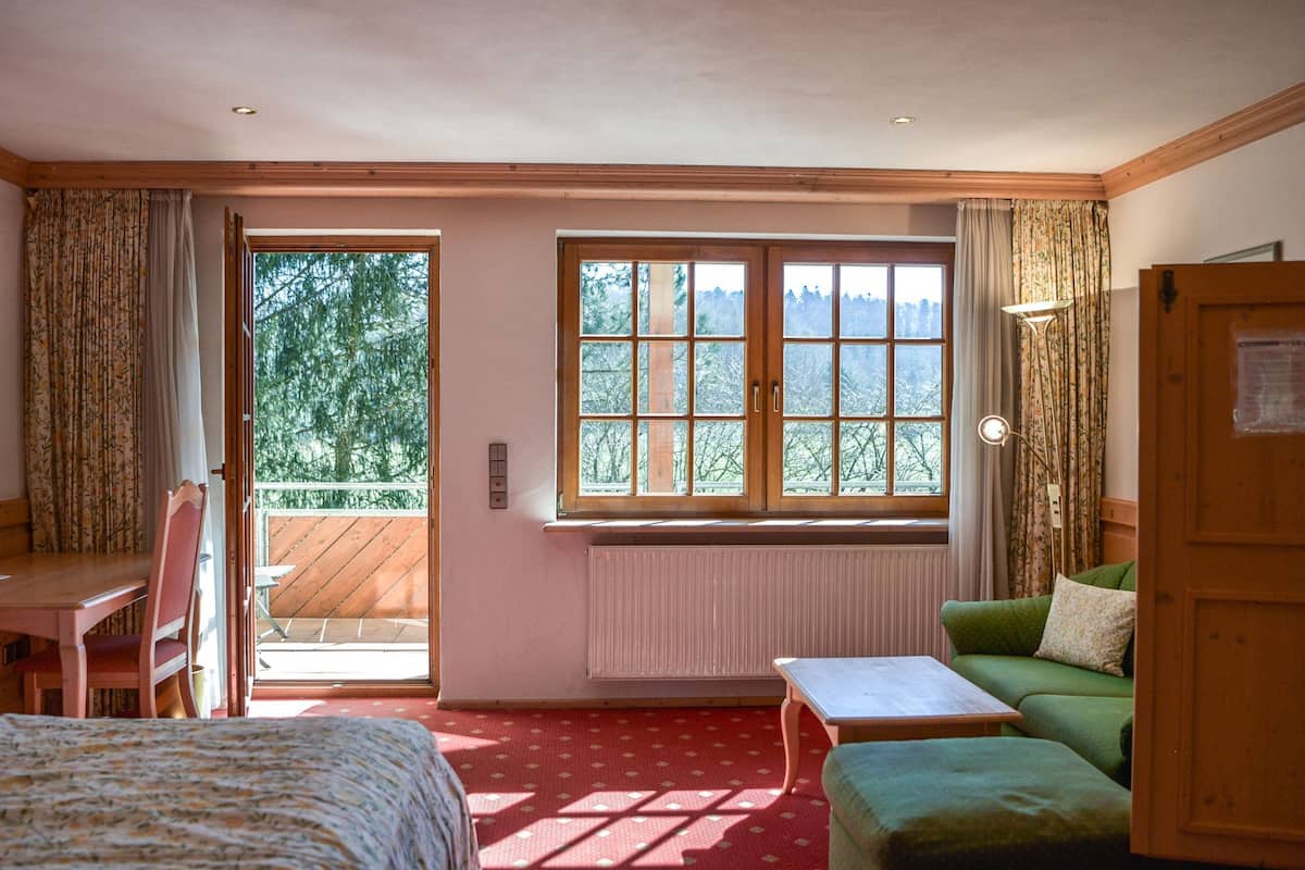 Una habitación con cama, sofá verde, una mesita y ventana con vista a árboles.