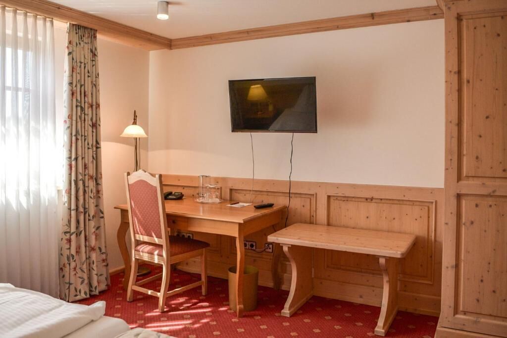 A hotel room with a wooden desk, a chair, a television mounted on the wall, and a bed with a red patterned carpet.