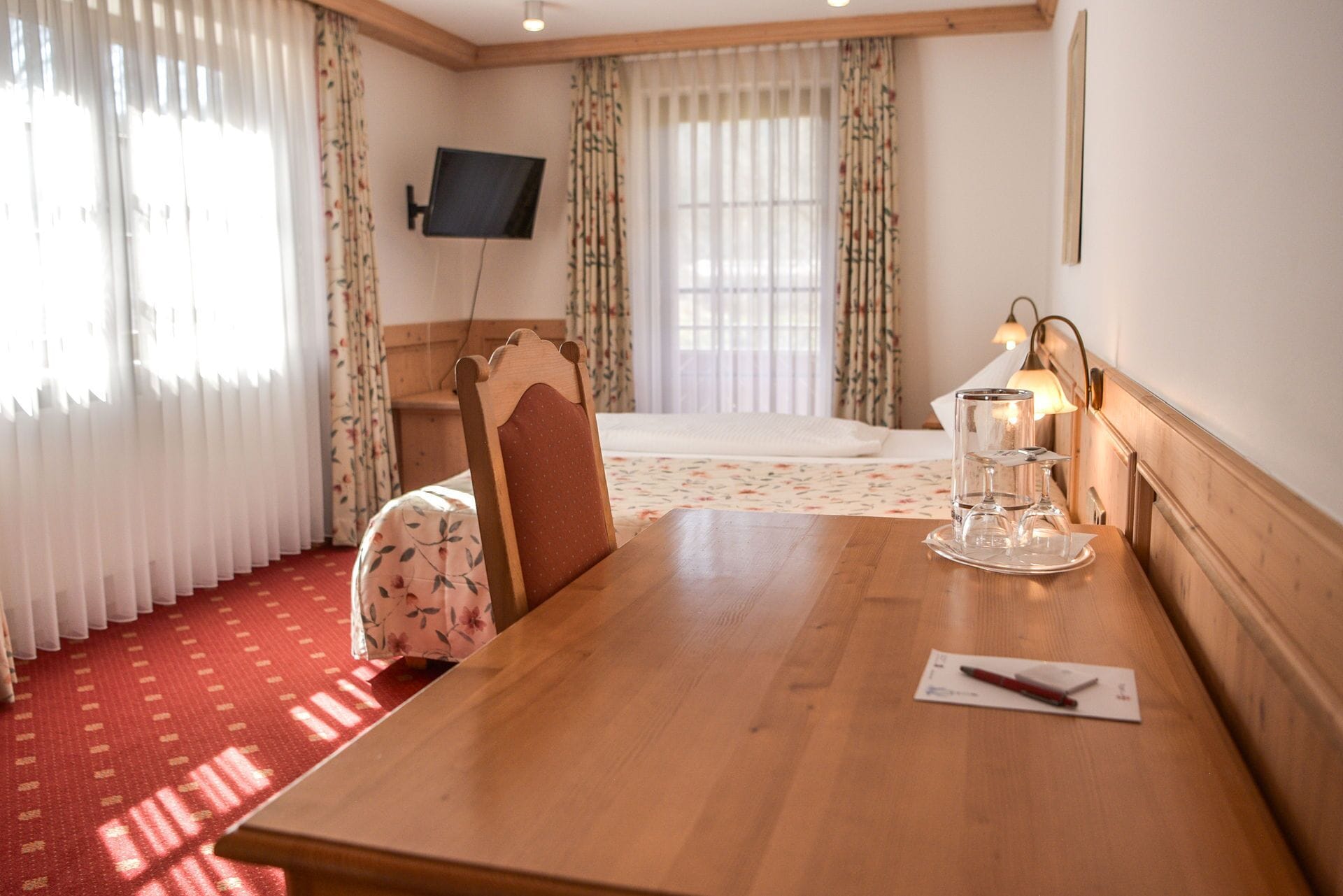 Habitación de hotel con mesa de comedor de madera, una silla, cama con colcha estampada, televisor montado en la pared y una lámpara de noche.