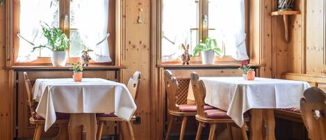 Un coin repas chaleureux, aménagé en bois, avec des tables et des chaises, des plantes en pot et une statue posée sur une étagère.