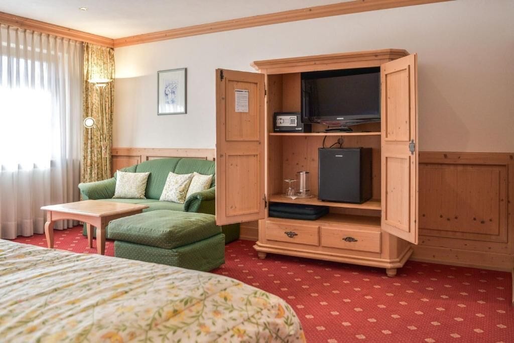 A hotel room with a bed, a green sofa, a wooden cabinet with a television and a small fridge, and a red carpet.