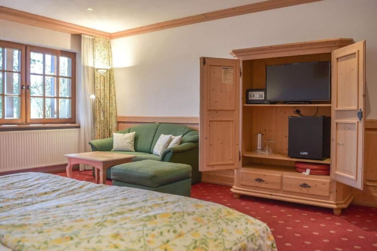 Una habitación con cama, sofá verde, mueble de madera con televisor y ventana con cortinas.