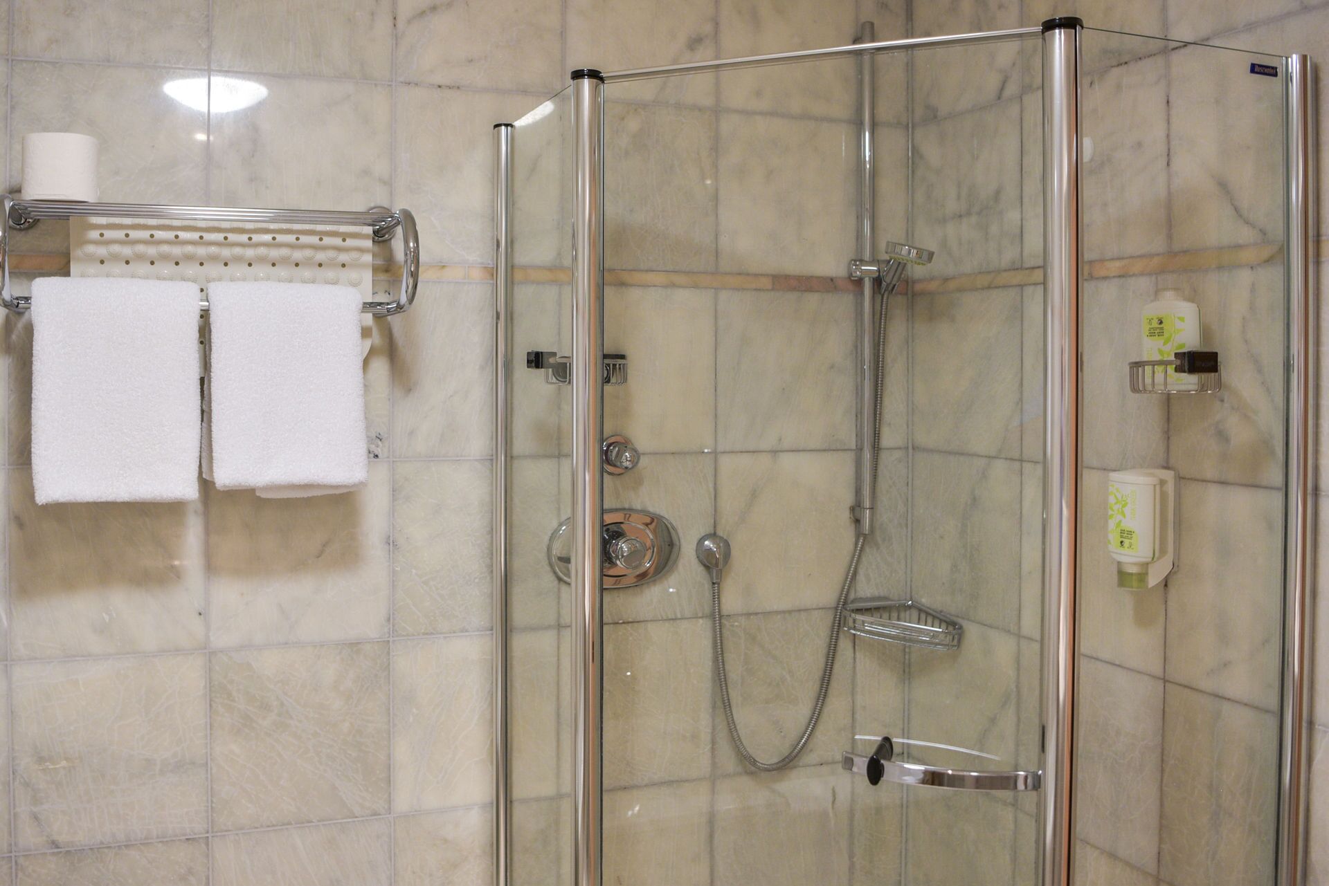A glass-enclosed shower stall with a towel rack holding two towels, a handheld showerhead, and a wall-mounted soap dispenser.