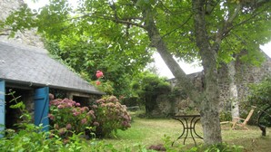 Ein rustikales Steinhaus mit geneigtem Dach, umgeben von üppigem Grün und blühenden Pflanzen.