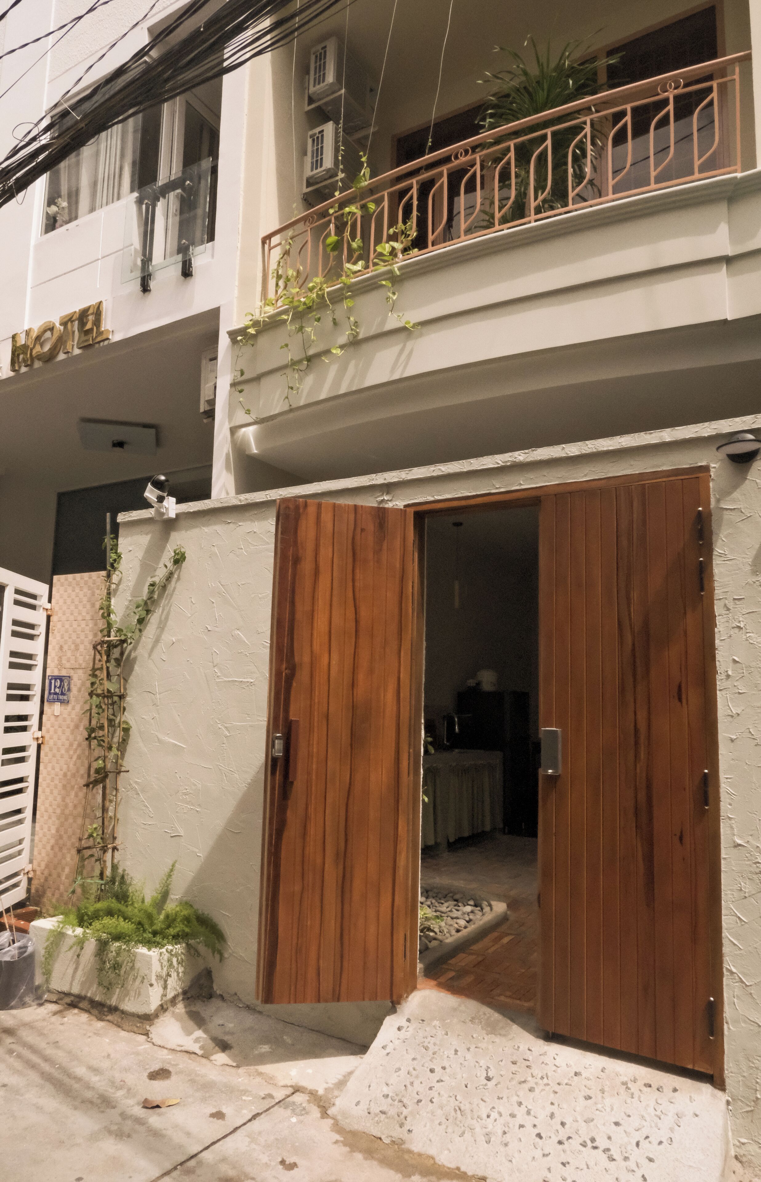 A hotel entrance with a wooden door, a small balcony with plants, and a visible room interior.
