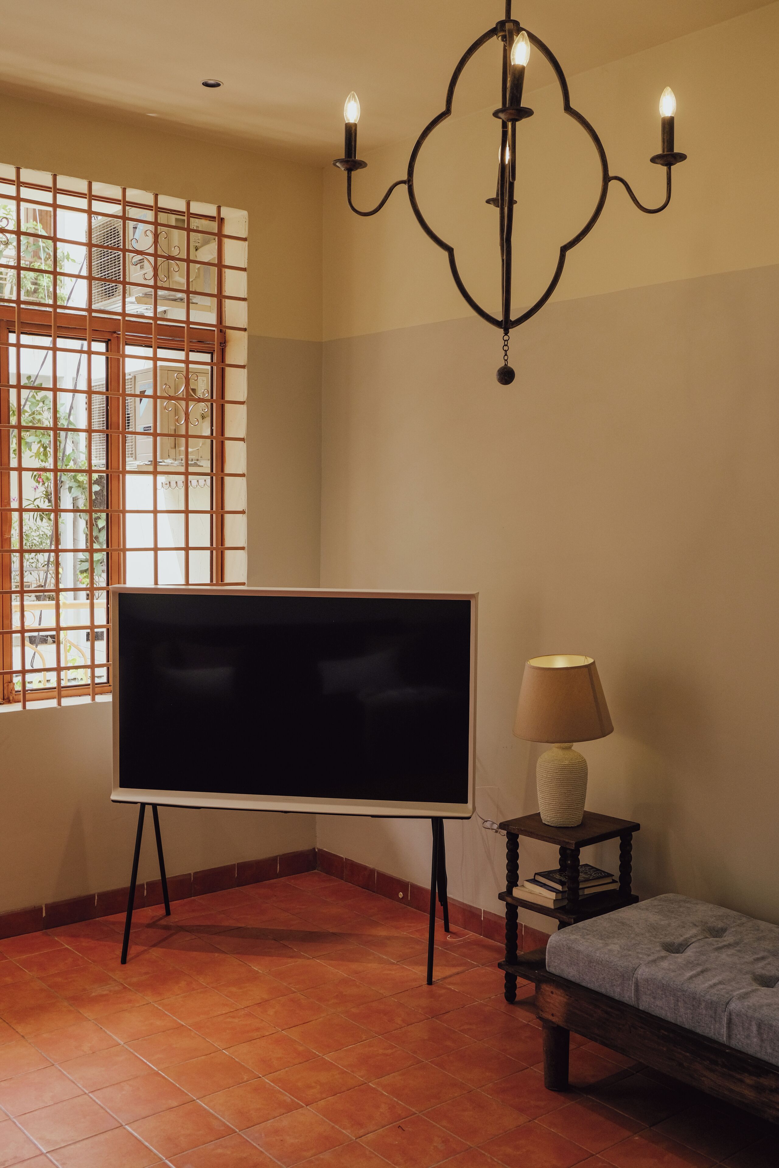 Una habitación con una televisión de pantalla plana grande sobre un mueble, una mesita con una lámpara, un banco y una lámpara decorativa montada en la pared.