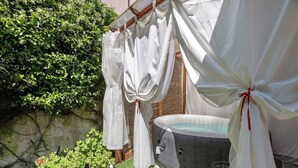 Una bañera de hidromasaje rodeada de cortinas blancas atadas con lazos rojos, con un fondo de follaje y una pared de hormigón.