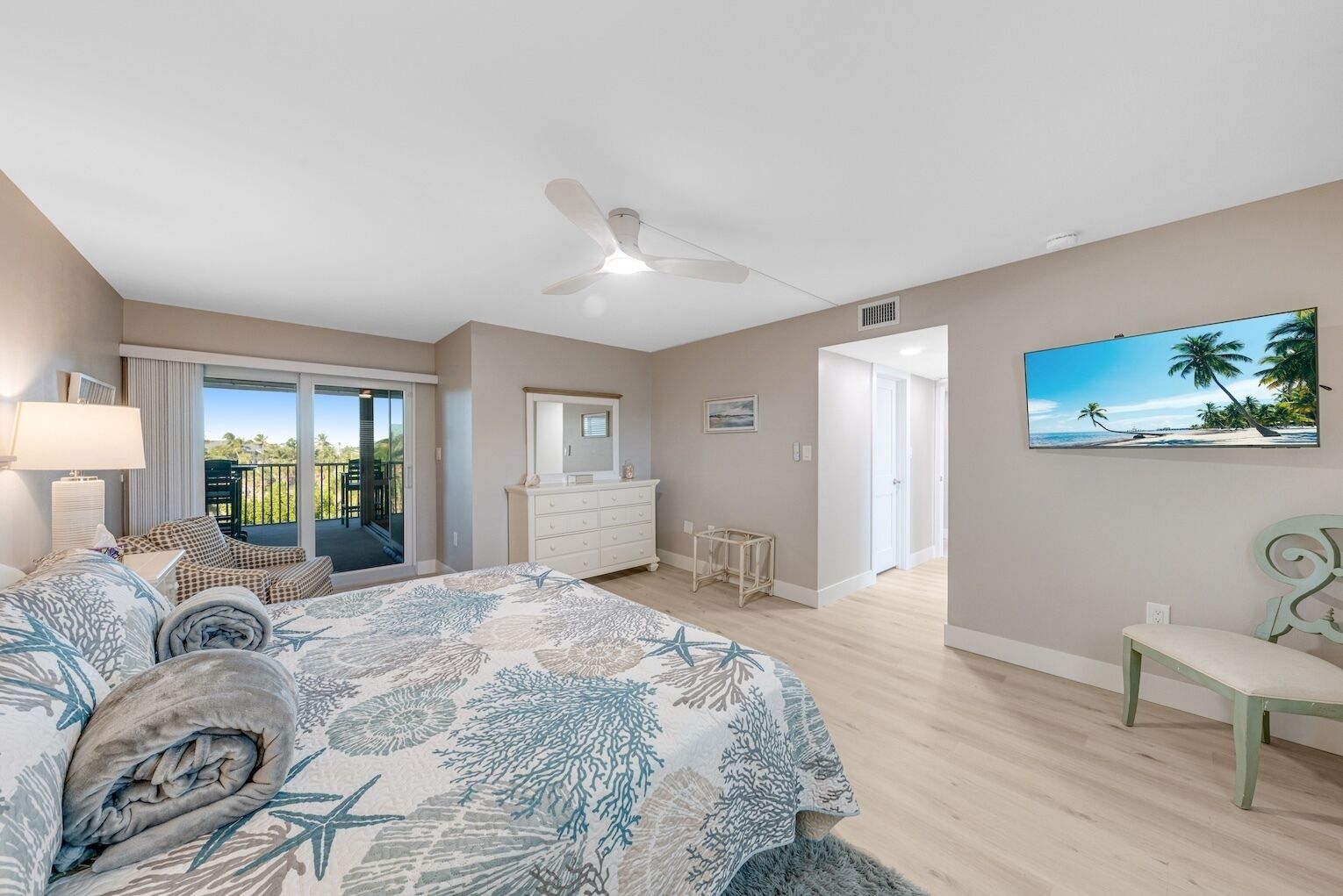 A bedroom with a bed, a dresser, a chair, a ceiling fan, and a painting of a beach.