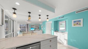 Una cucina moderna con isola centrale, mobili bianchi e pareti color verde acqua. Sono presenti luci a sospensione, una zona pranzo con tavolo e sedie bianche, e vista sull'area soggiorno attraverso una porta aperta.