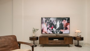A modern living room with a flat-screen TV mounted on a wooden TV stand, a brown leather sofa, a small side table with a plant, and a floor lamp. - Tamundi Embassy - Diplomatic Quarter - West Lake Hanoi (Hanoi)