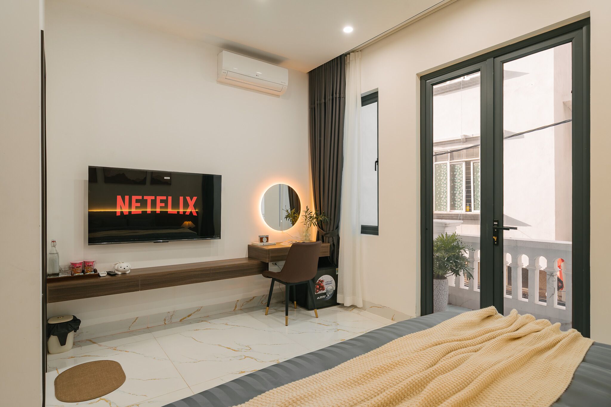 Ein modernes Schlafzimmer mit Fernseher, der 'NETFLIX' zeigt, einem Schreibtisch mit Spiegel, einem Bett mit Decke und Blick auf einen Balkon.