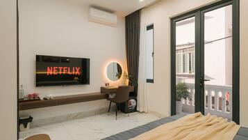 Ein modernes Schlafzimmer mit Fernseher, der 'NETFLIX' zeigt, einem Schreibtisch mit Spiegel, einem Bett mit Decke und Blick auf einen Balkon.