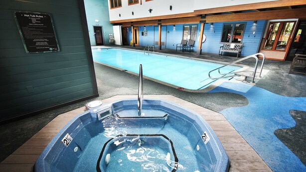 Un bain à remous avec un escalier en métal, une piscine avec une terrasse bleue et un bâtiment doté de grandes fenêtres et d’un mur extérieur bleu.