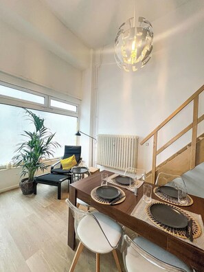 Et moderne spiseområde med et træbord dækket til to, transparente glasstole og et hængende lysarmatur.