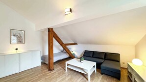 Camera da letto moderna mansardata, con soffitto spiovente, struttura in travi di legno, letto bianco, divano scuro, tavolino da caffè bianco e lampada da comodino.