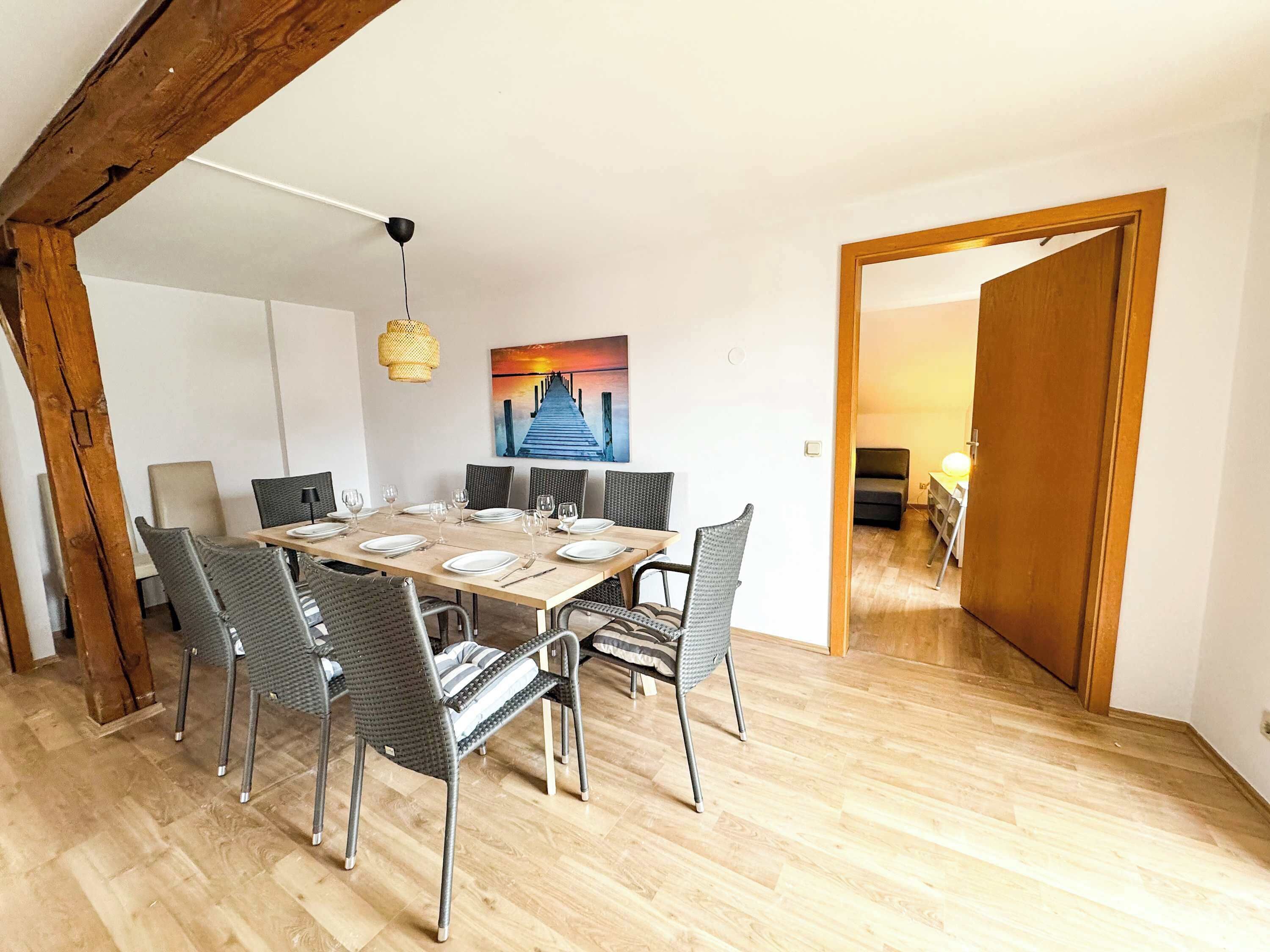 A dining area with a wooden table set for six, surrounded by wicker chairs. A painting of a pier is on the wall, and a wooden door leads to another room.
