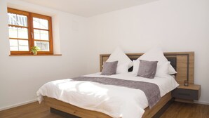Un dormitorio con cama de madera, ropa de cama blanca, una mesita de noche y una pequeña, además de una ventana con una planta.