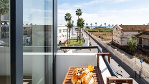 Un balcón con una mesa dispuesta con cruasanes y jugo de naranja, con vistas a una zona costera con palmeras y un edificio histórico.