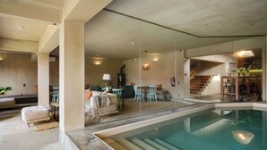 Ein moderner Innenpool mit großen Glaswänden, eine Lounge-Bereich mit Sofas und Sesseln sowie eine Treppe, die in eine obere Ebene führt.