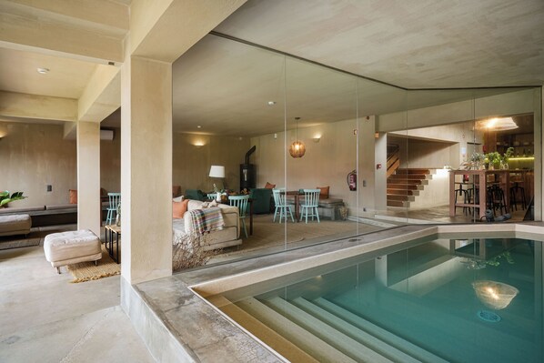 Ein moderner Innenpool mit großen Glaswänden, eine Lounge-Bereich mit Sofas und Sesseln sowie eine Treppe, die in eine obere Ebene führt.