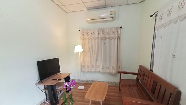 Een woonkamer met een televisie, een houten bankje, een bijzettafeltje, een potplant, een lamp en een raam met gordijnen.
