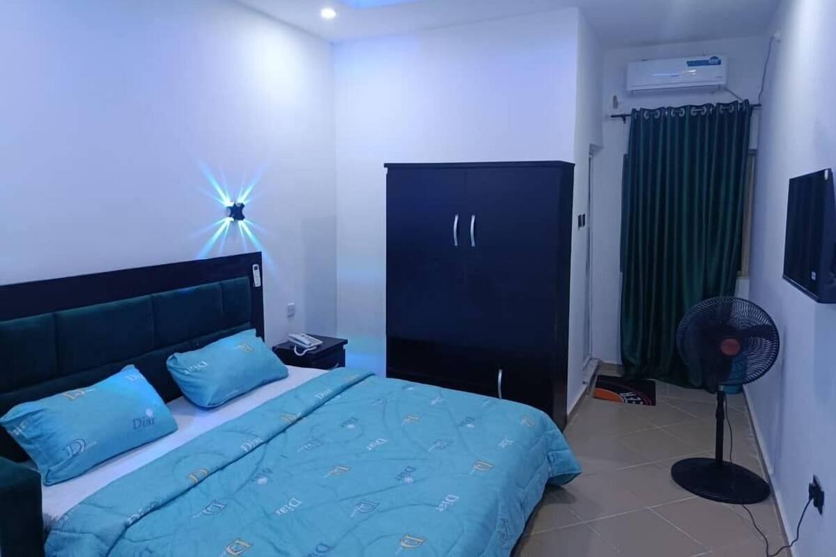 A bedroom with a bed, a wardrobe, a desk, a standing fan, and an air conditioning unit.