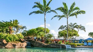 Ein klares Schwimmbecken mit Wasserfall, Palmen und einer Liegefläche mit Sonnenschirmen.