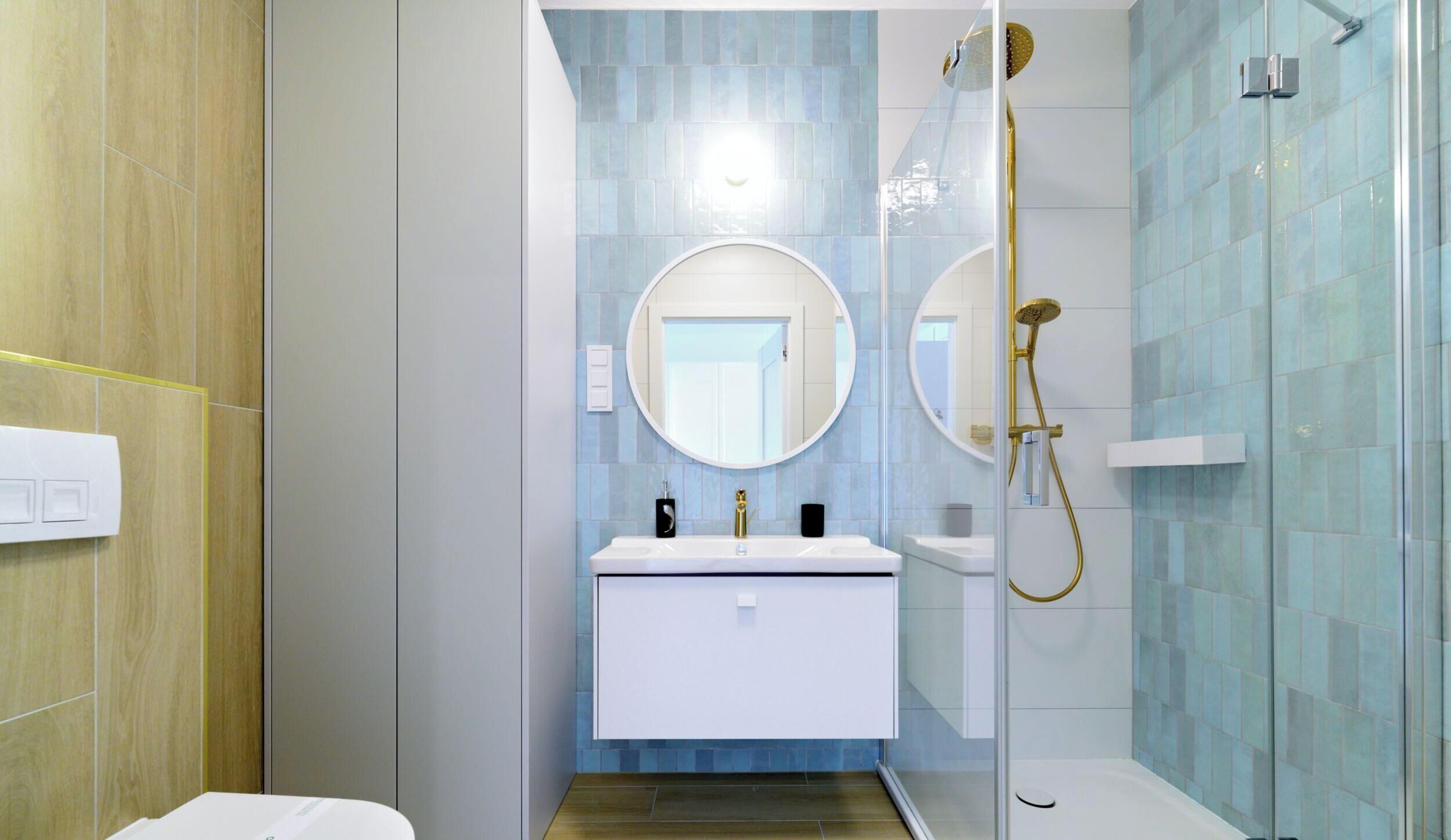 A modern bathroom with a glass-enclosed shower, a white vanity with a gold faucet, and a round mirror above the sink.