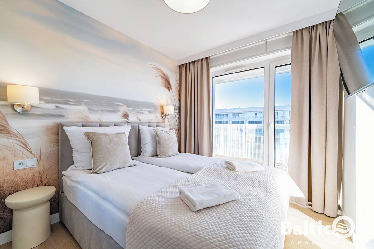 Una camera d'albergo con un letto grande, un comodino, una lampada a parete e vista sulla città attraverso ampie finestre.