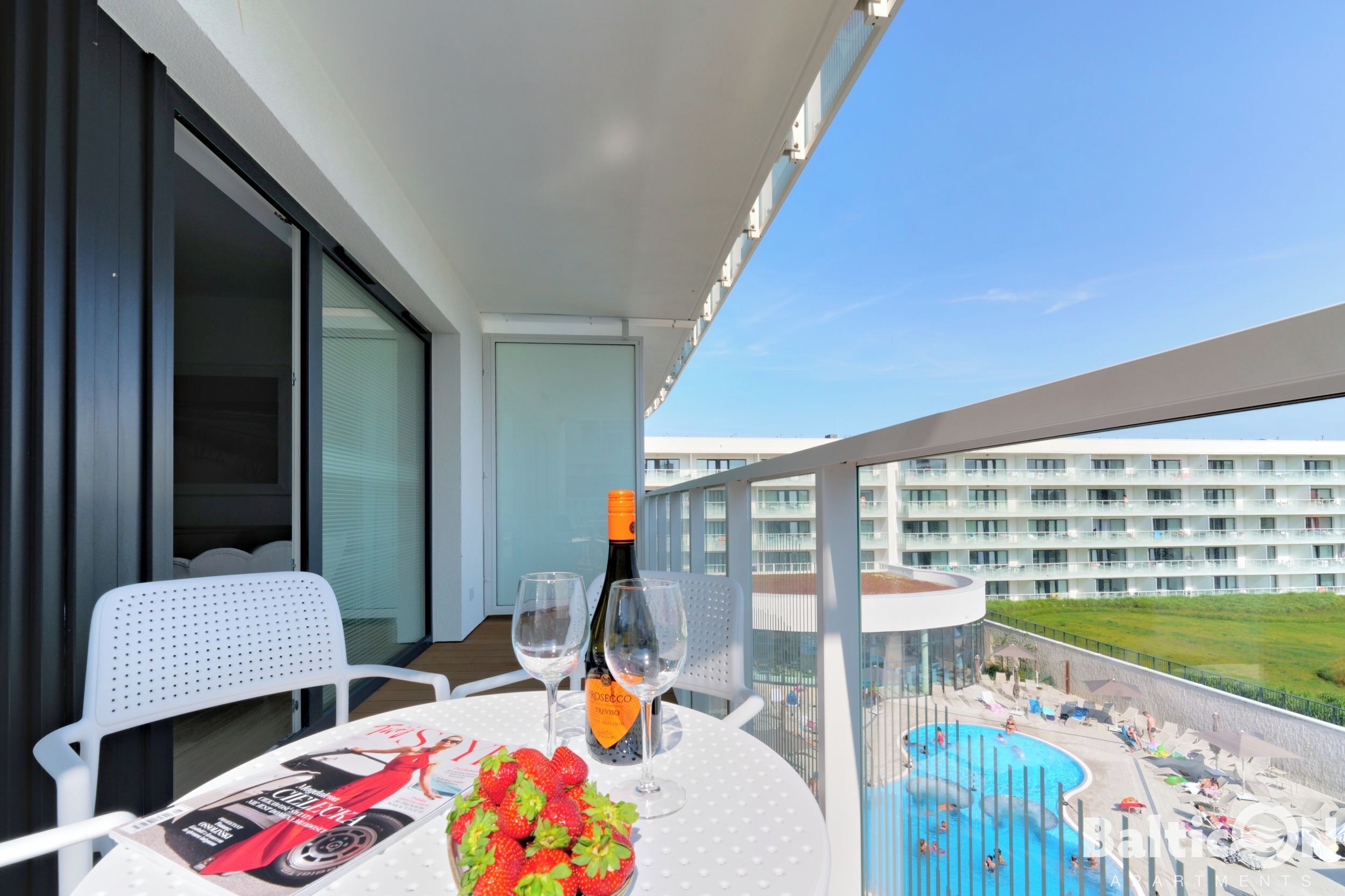 Un balcon avec une table, deux chaises, une bouteille de vin rosé, deux verres, un saladier de fraises et un magazine.