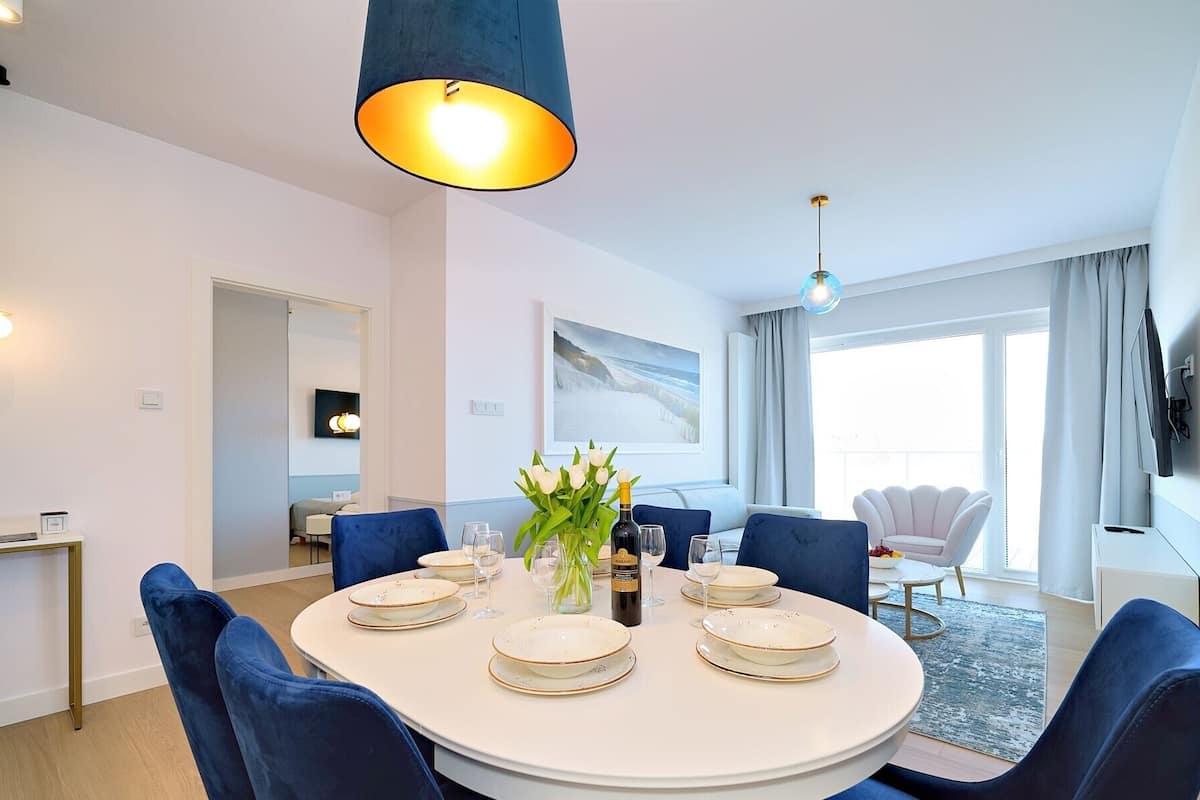 Un'elegante zona pranzo con un tavolo rotondo apparecchiato per quattro persone, sedie rivestite in blu, un vaso con fiori e una bottiglia di vino.