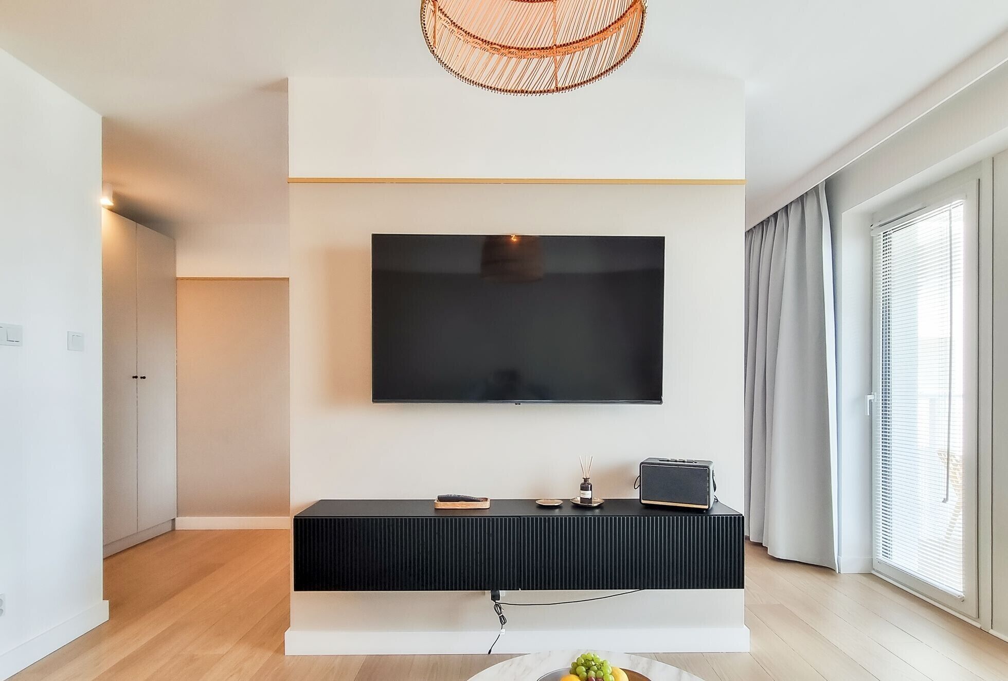 A modern living room with a flat-screen TV mounted on a white wall, a black console table below, and a woven pendant light above.
