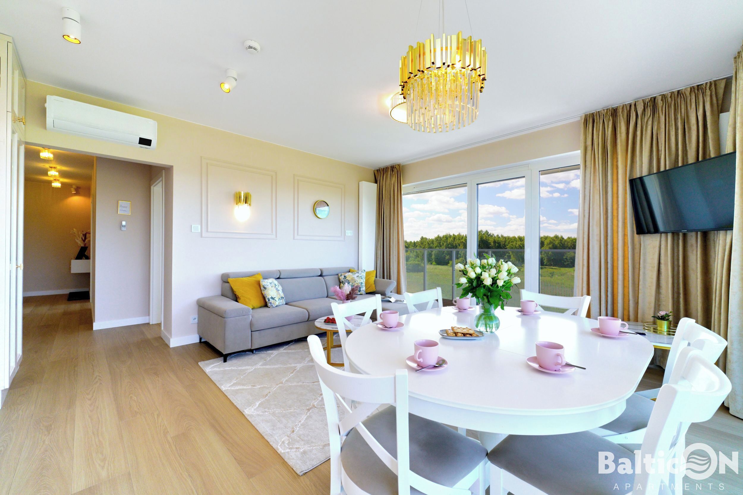 A modern living room with a dining area, a grey sofa with yellow cushions, a chandelier, and a TV mounted on the wall.