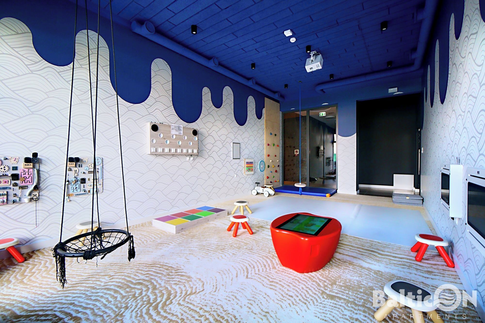 Zona infantil con un columpio colgante, alfombras de juego coloridas y un sillón de tierno rojo.