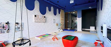 Zona infantil con un columpio colgante, alfombras de juego coloridas y un sillón de tierno rojo.