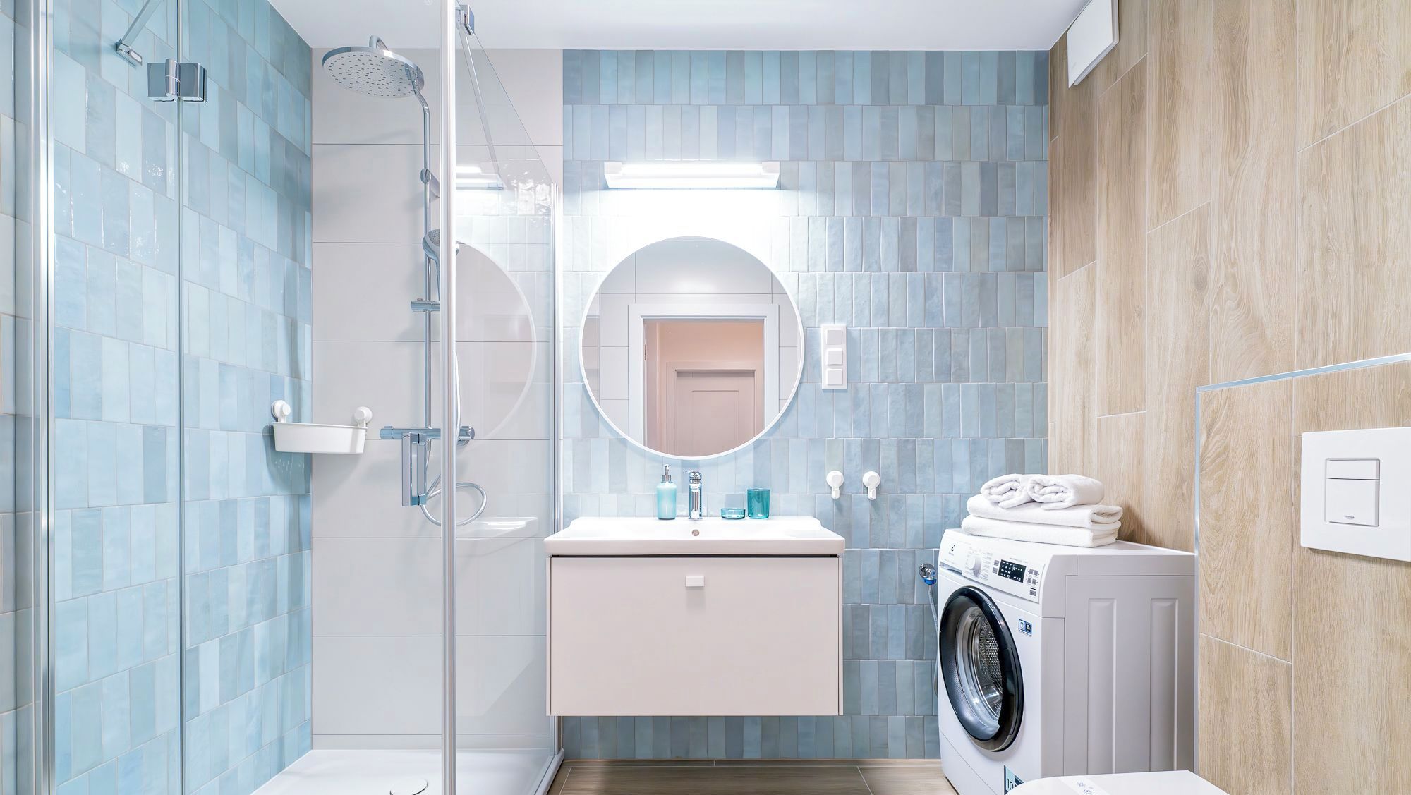 Un baño moderno con cabina de ducha de vidrio, un espejo redondo, un lavabo con encimera blanca y una lavadora empotrada.