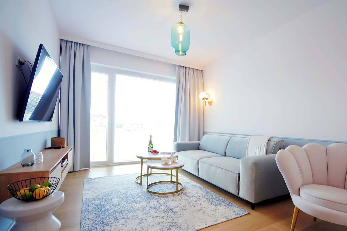 Una sala de estar moderna con un sofá, una mesa de centro redonda, un sillón rosa, un televisor de pantalla plana montado en la pared y un ventanal con cortinas.