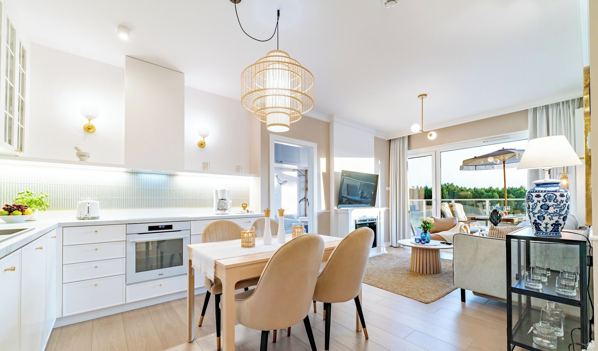 A modern kitchen with white cabinetry, a dining area with a table and chairs, and a living area with a sofa and a TV.